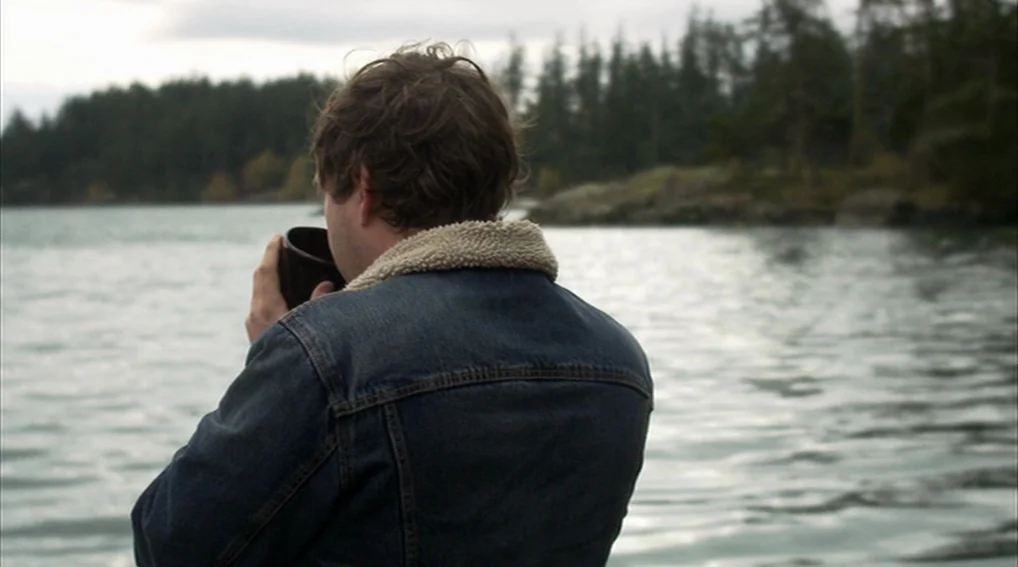 Your Sister’s Sister (2011) movie still: medium — A man stands on a rocky shore beside a calm, gray lake with dense trees. The man is wearing a…