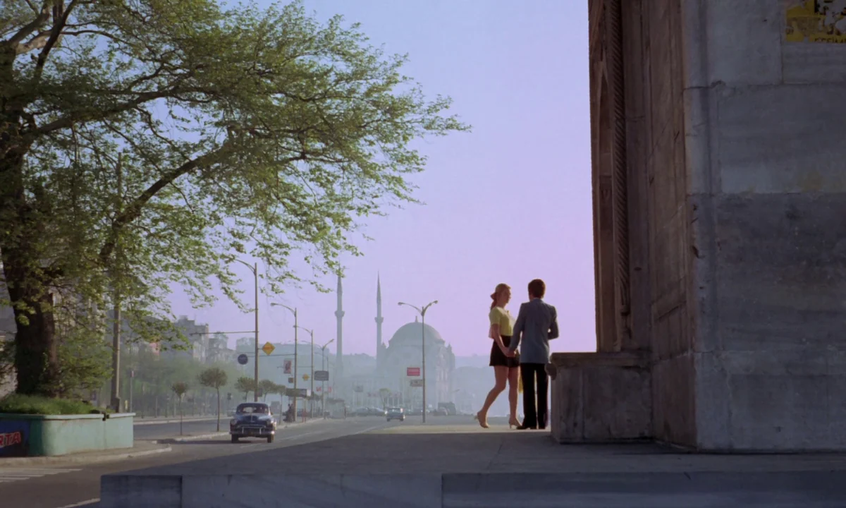Vampyros Lesbos (1971) movie still: wide — A wide shot shows a couple standing on a stone ledge in an urban setting. A large mosque with two…