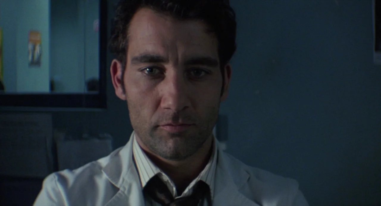 Closer (2004) movie still: closeup — A dark, dimly lit office space with a blurred wall poster visible in background. A man in a light pinstriped…