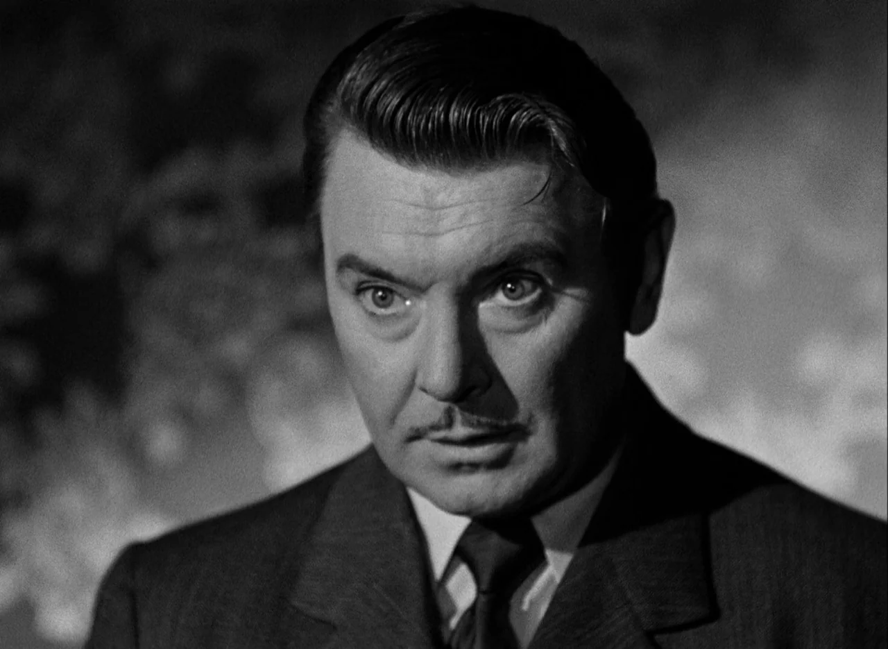 The Spiral Staircase (1946) movie still: closeup — A dimly lit room with textured, shadowed surfaces in the background. A man stares forward with an…