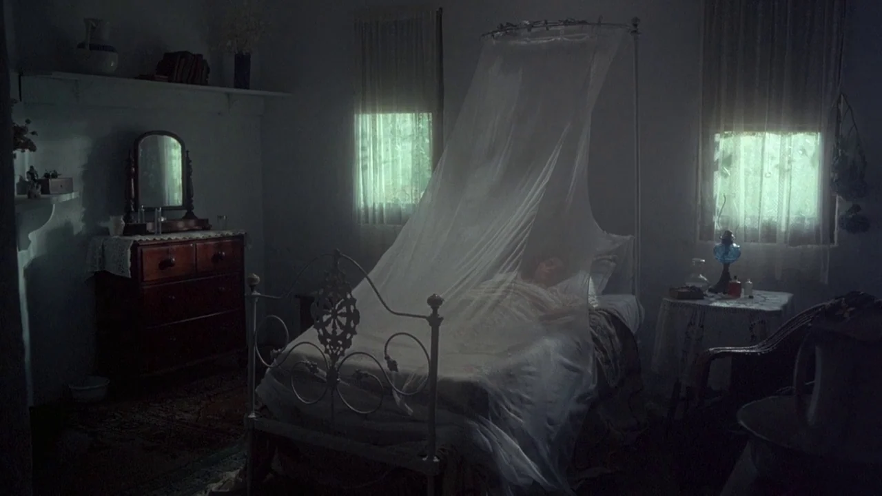 Picnic at Hanging Rock (1975) movie still: wide — A dimly lit room contains an iron bed draped in white mosquito netting. A person rests under the…