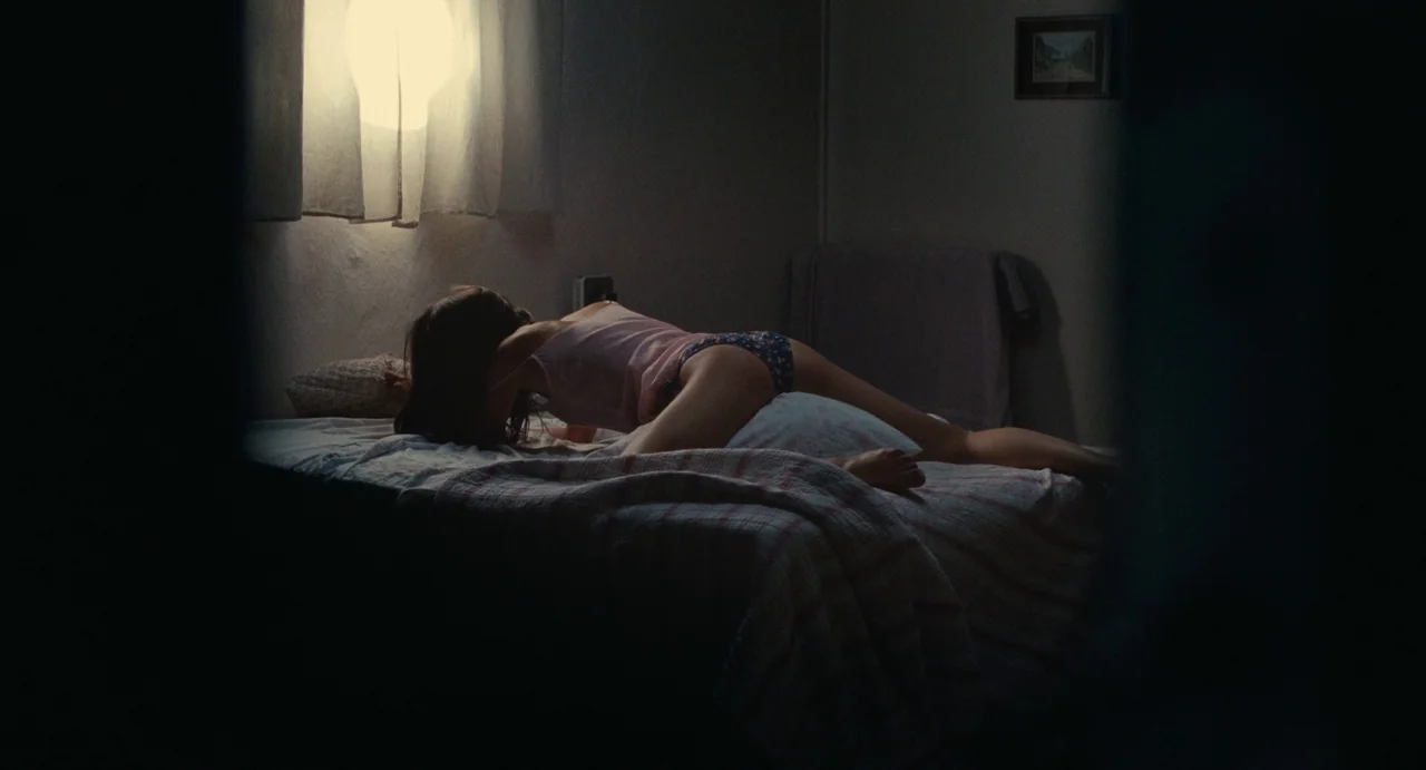 Young & Beautiful (2013) movie still: medium — A dimly lit room contains a bed with a light-colored, patterned blanket. A person lies on their…