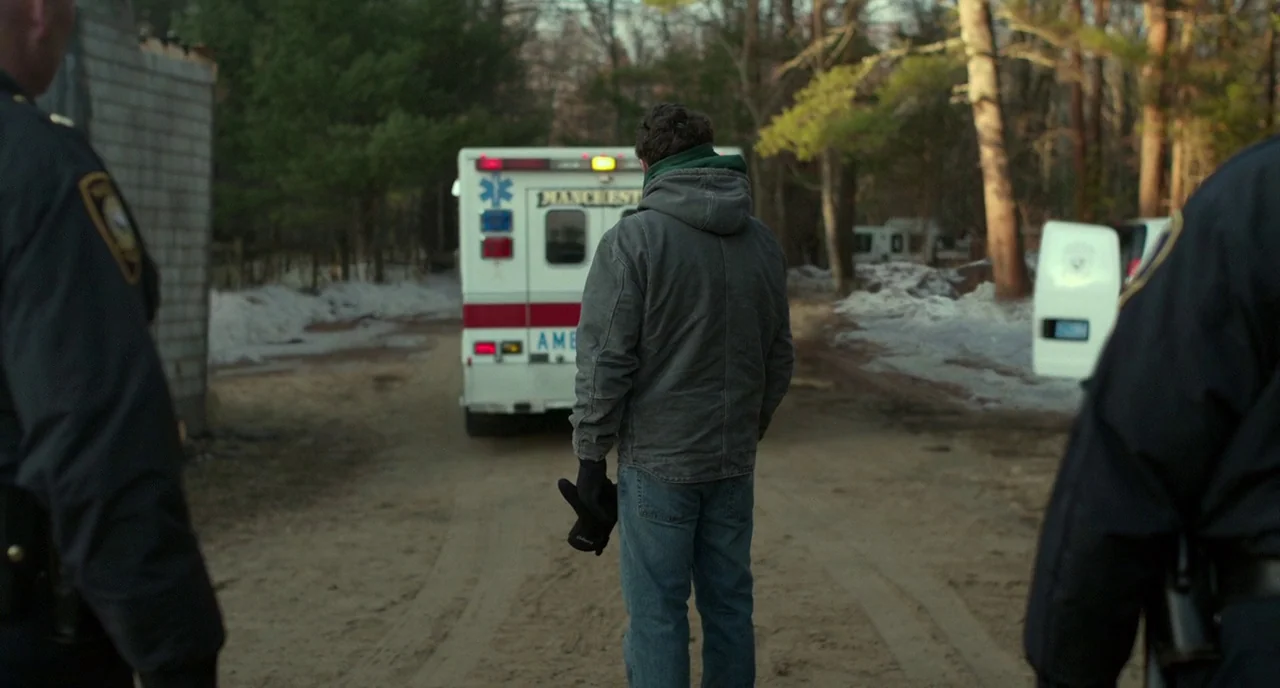 Manchester By The Sea (2016) movie still: medium — A dirt road in a wooded area contains patches of melting snow. A white ambulance with lights on…