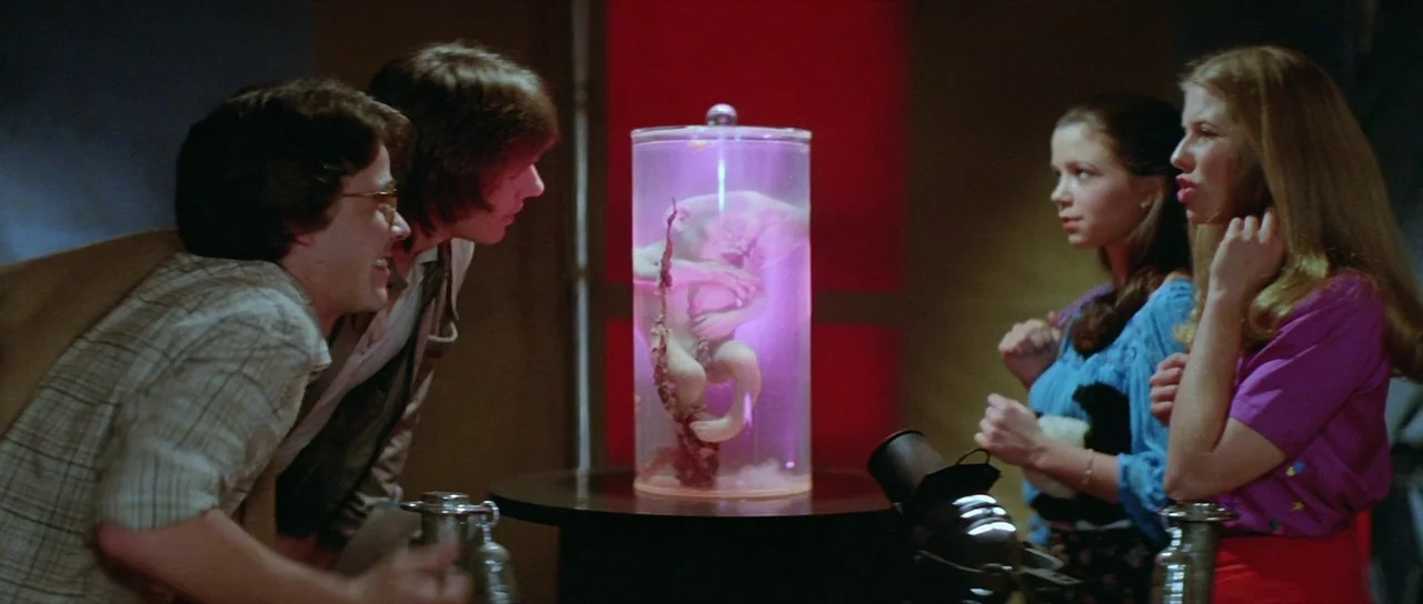 The Funhouse (1981) movie still: medium — An exhibit room features a large cylindrical glass tank illuminated with purple liquid inside. Four young…