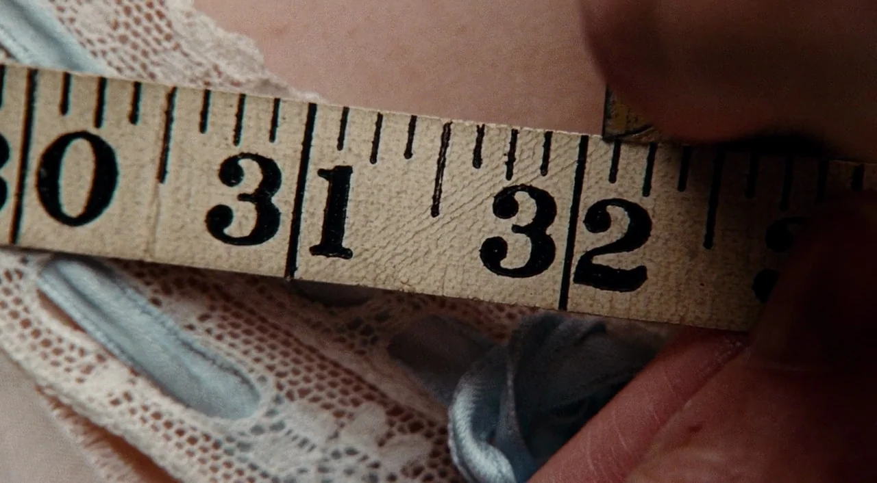 Phantom Thread (2017) movie still: closeup — A close view of a yellow measuring tape pressed against light lace fabric. The digits thirty-one and…