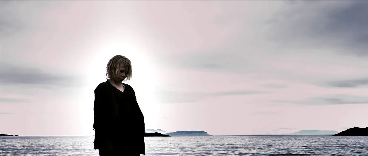 Valhalla Rising (2009) movie still: medium — A person stands on a rocky shoreline beneath a bright, overcast sky. The figure is silhouetted against…