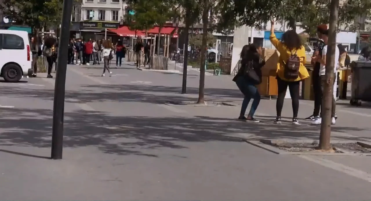 A Dramatic Film (2019) movie still: fullbody — An urban plaza paved with grey concrete features young pedestrians and scattered trees. Several…
