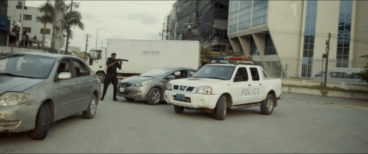 Saving Onome (2024) movie still: wide — An asphalt road intersection features a white police pickup truck and several civilian cars. A uniformed…