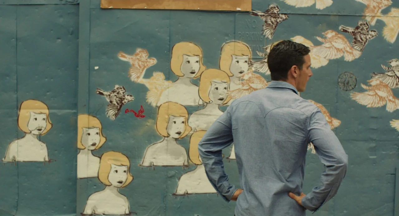 Cymbeline (2014) movie still: medium — A blue construction fence serves as a canvas for a white-and-yellow illustrative mural. A man in a blue shirt…