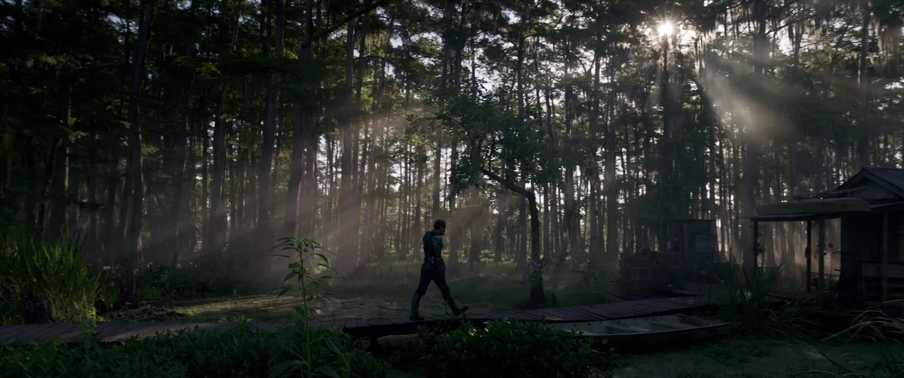 Captain Marvel (2019) movie still: wide — A dense cypress swamp features thick trees draped with hanging moss and murky water. A single man walks…