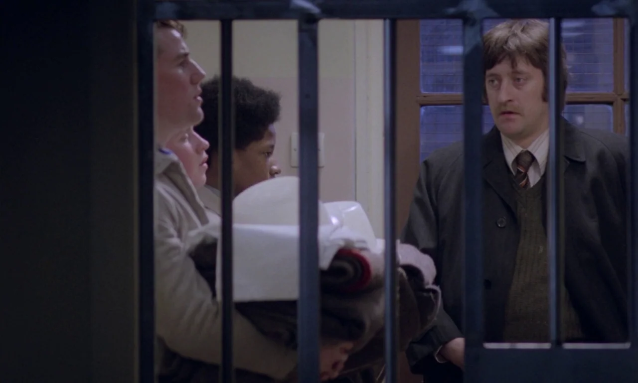 Scum (1979) movie still: medium — The scene shows a group inside a secure area with vertical metal bars visible. A man in a dark coat stands outside…
