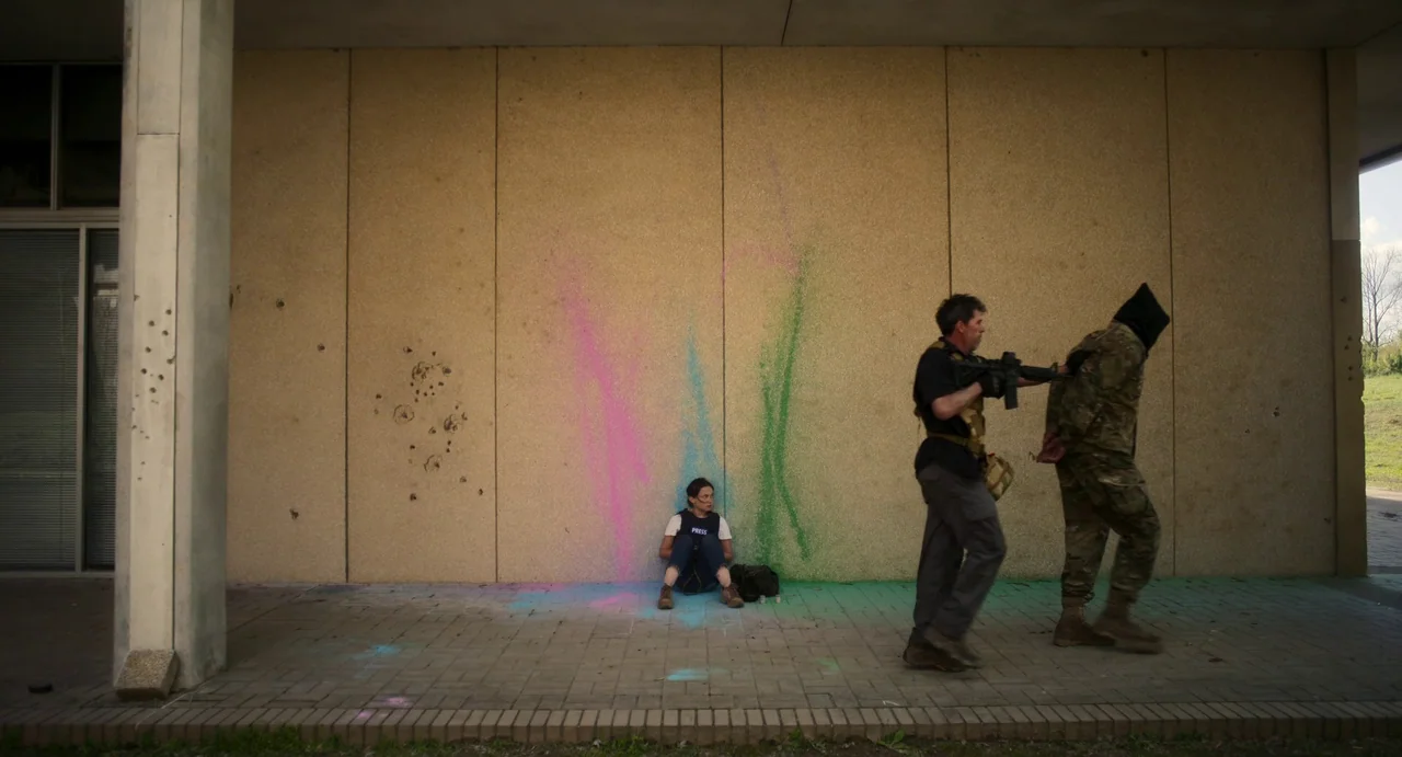 Civil War (2024) movie still: wide — A woman sits on a paved area against a wall splattered with colored paint. An armed man leads a hooded prisoner…