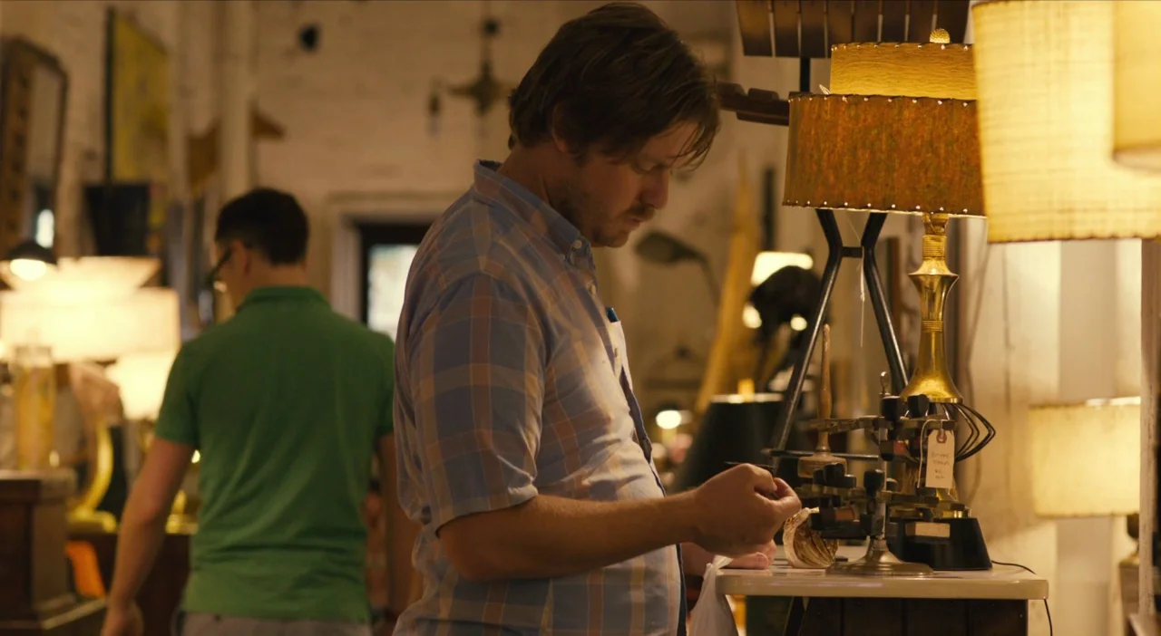 The Comedy (2012) movie still: medium — A cluttered shop interior contains numerous vintage lamps and antique inventory items on display. A man…