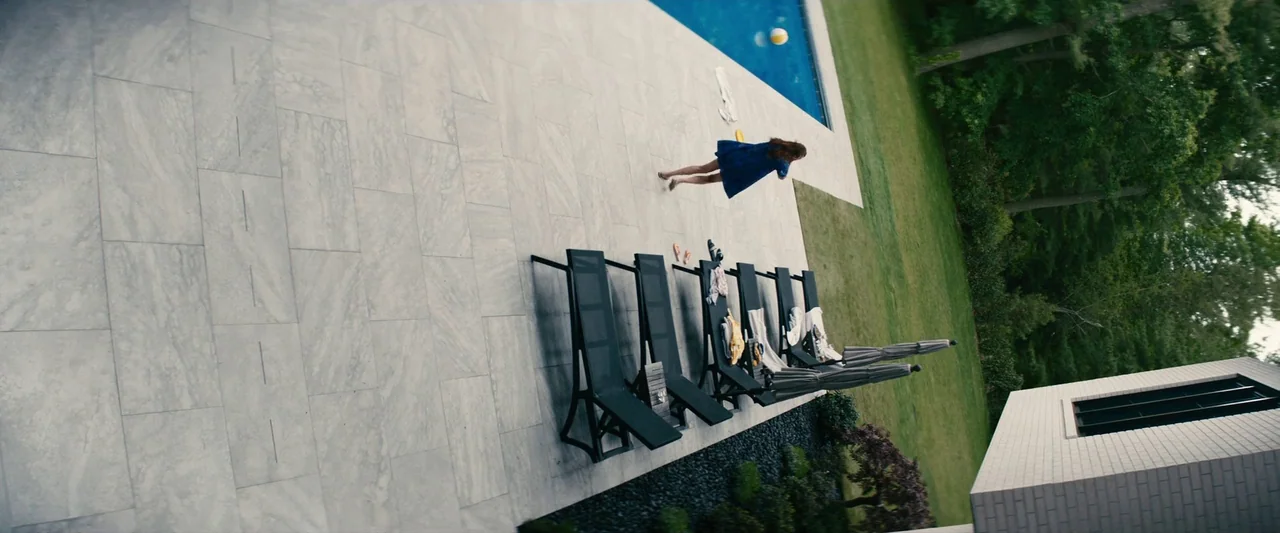 Leave The World Behind (2023) movie still: wide — A modern stone patio adjacent to a swimming pool with lounge chairs scattered around. A woman…