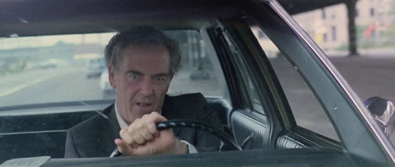The New York Ripper (1982) movie still: medium — A man drives a car through an urban street during the daytime. The car interior is visible with the…