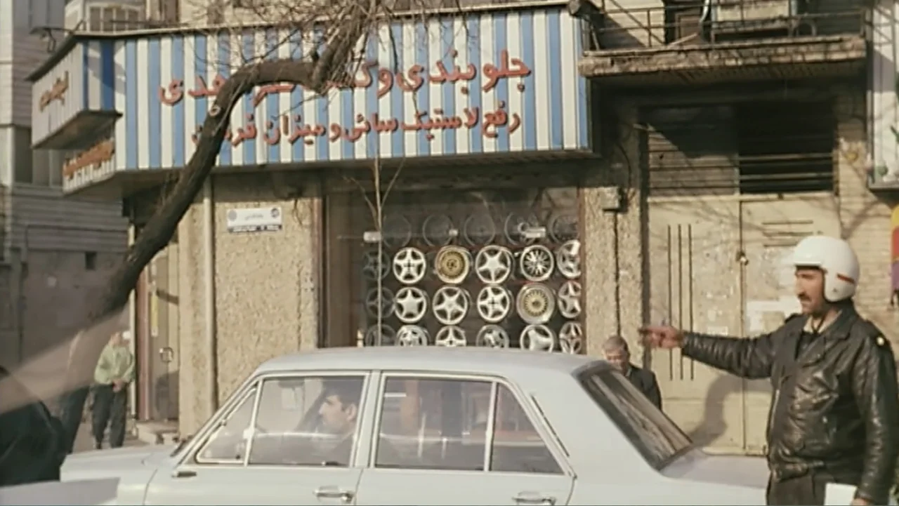 The Mirror (2014) movie still: medium — An exterior street scene shows a shopfront displaying various car rims behind a glass window. A man in a…