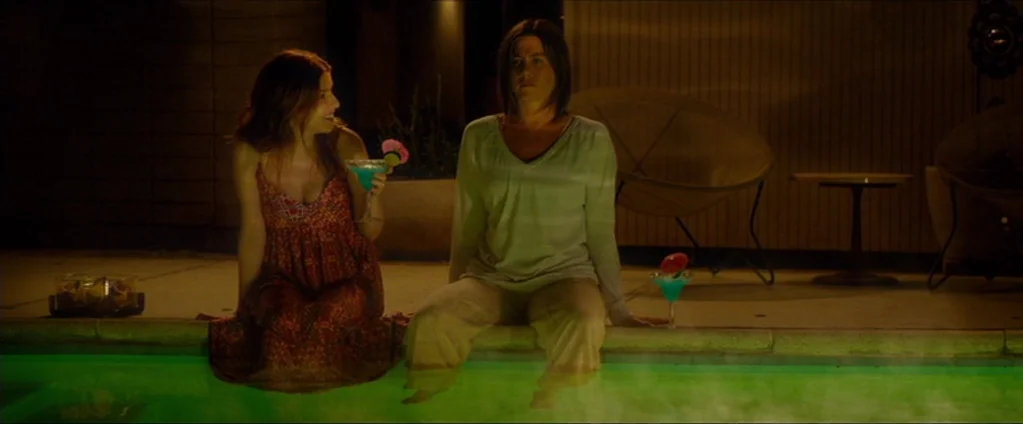 Cake (2014) movie still: medium — Two women sit at the tiled edge of an illuminated pool at night. They hold blue cocktails while positioned in…