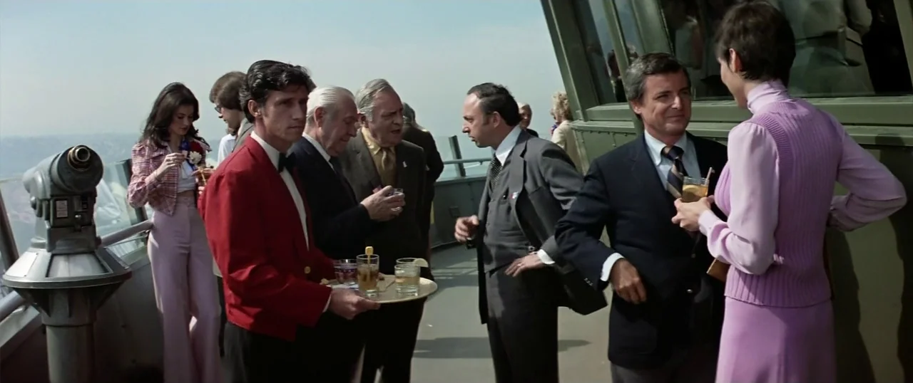The Parallax View (1974) movie still: medium — A group of people socialize on a metal-railed observation deck during a sunny day. A man in a red…