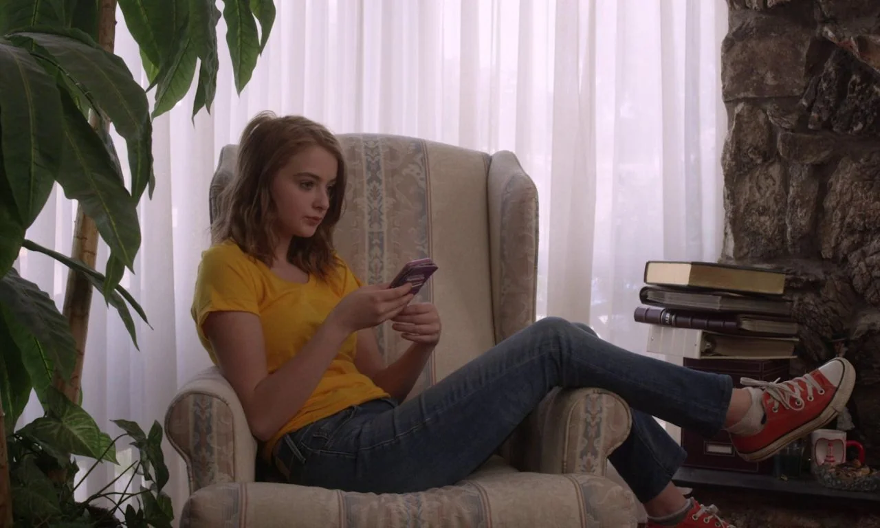 Ham on Rye (2019) movie still: medium — A woman sits in a patterned armchair in front of floor-to-ceiling white curtains. She holds a purple…