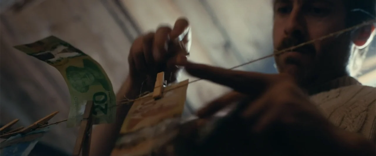A Small Fortune (2021) movie still: closeup — A dimly lit room contains a string line stretched across the foreground. A man attaches paper currency…
