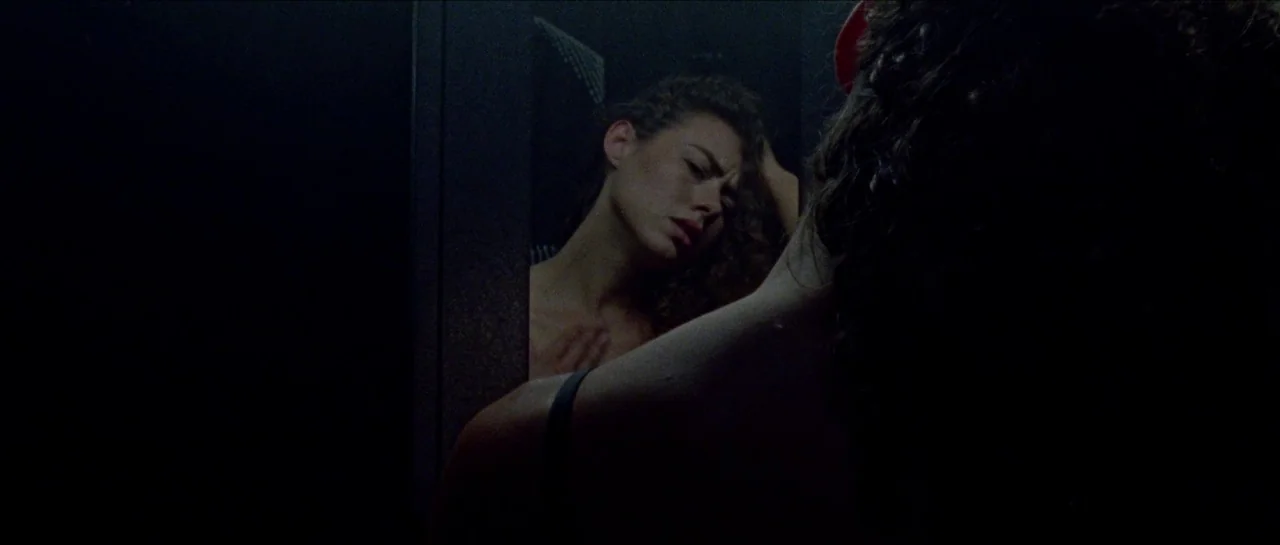 Bliss movie still: medium — A dimly lit room containing a reflective surface in the dark. A woman looks at her own reflection while touching her…