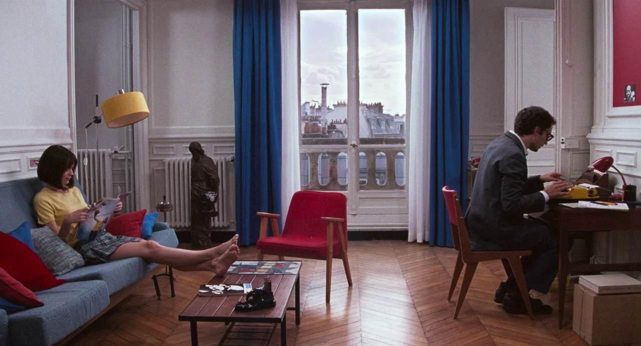 Godard Mon Amour (2015) movie still: wide — A Parisian apartment features a herringbone wood floor and white walls with colorful furniture. A woman…