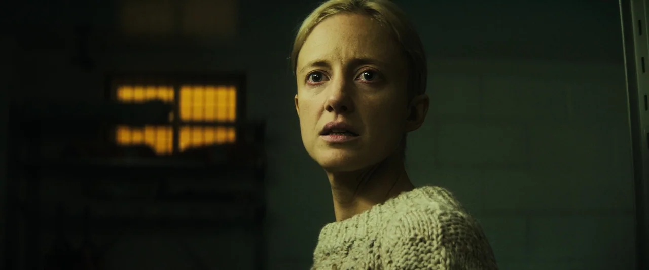 The Grudge (2020) movie still: closeup — A dark space features a small, glowing window with vertical bars. A woman with a concerned expression gazes…
