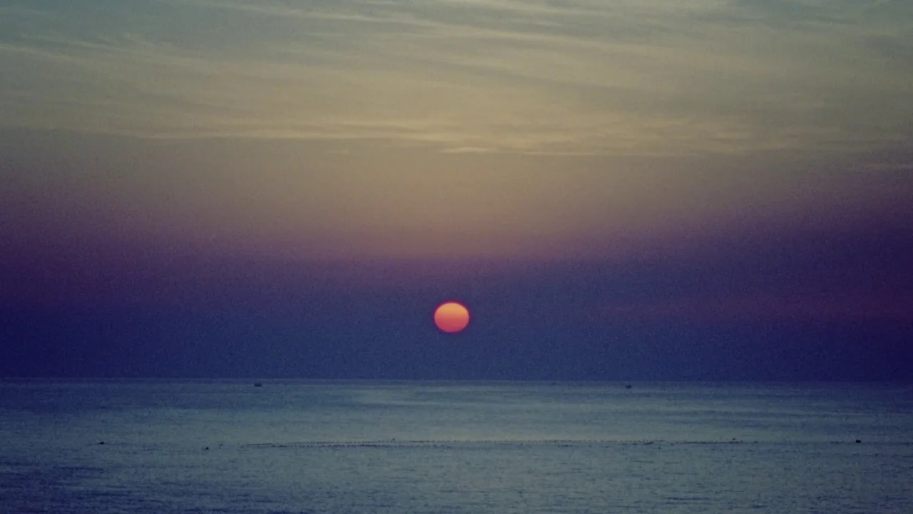 Riceboy Sleeps (2022) movie still: establishing — A vast calm ocean stretches out beneath a gradient sky at twilight. The round orange sun sits just…