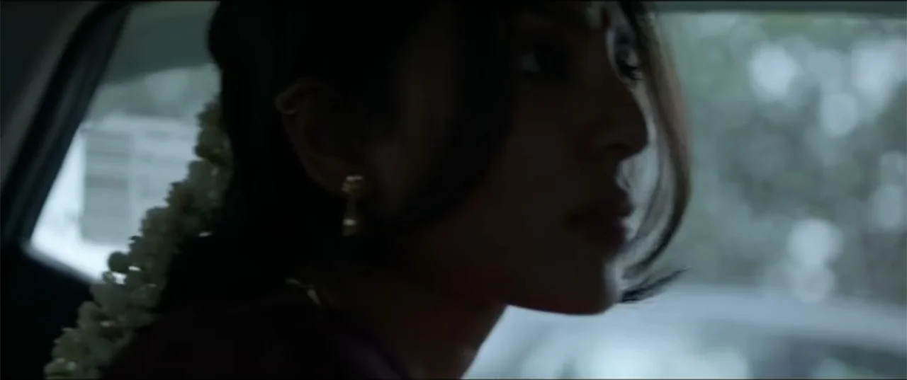 Cheekatilo (2026) movie still: closeup — An interior of a car with a blurry view of foliage through the window. A woman with jasmine flowers in her…