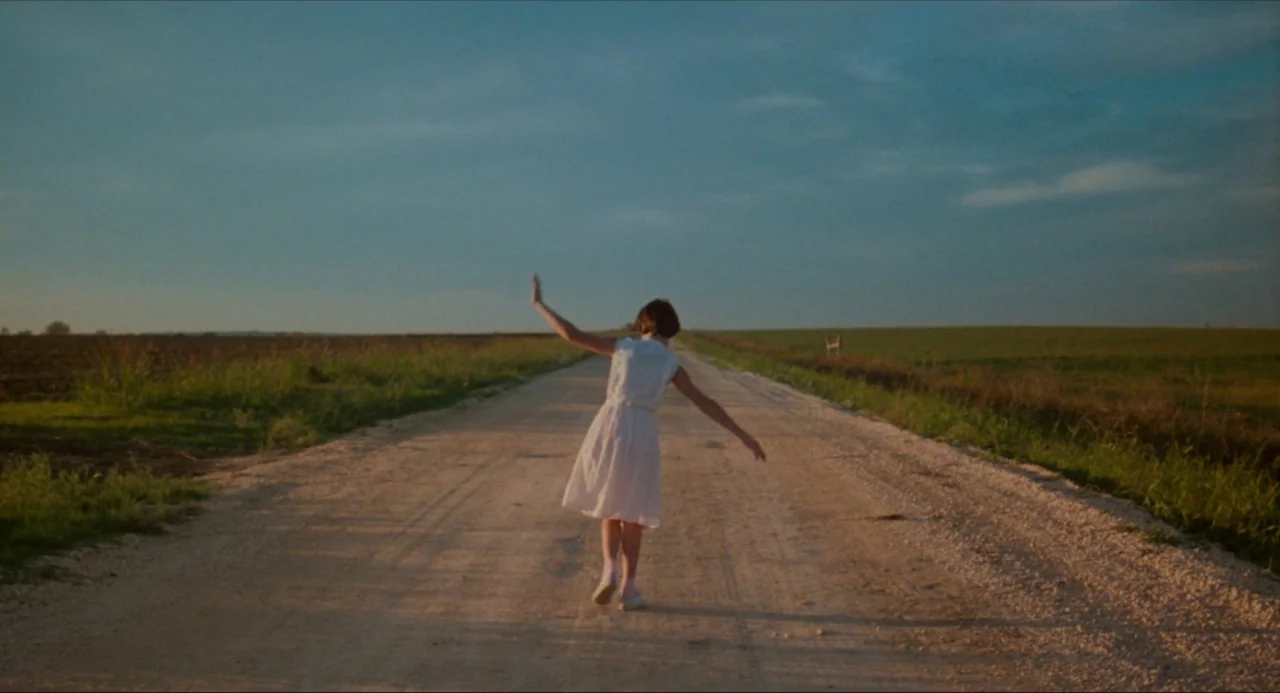 True Stories (1986) movie still: fullbody — A long, straight gravel path extends through vast, flat grassy fields under a sky. A young girl in a…