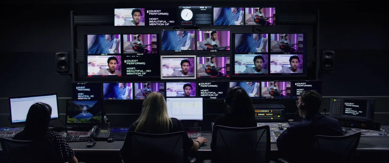 Yesterday (2019) movie still: wide — A dimly lit production control room features a large video wall displaying various feeds. Four staff members…