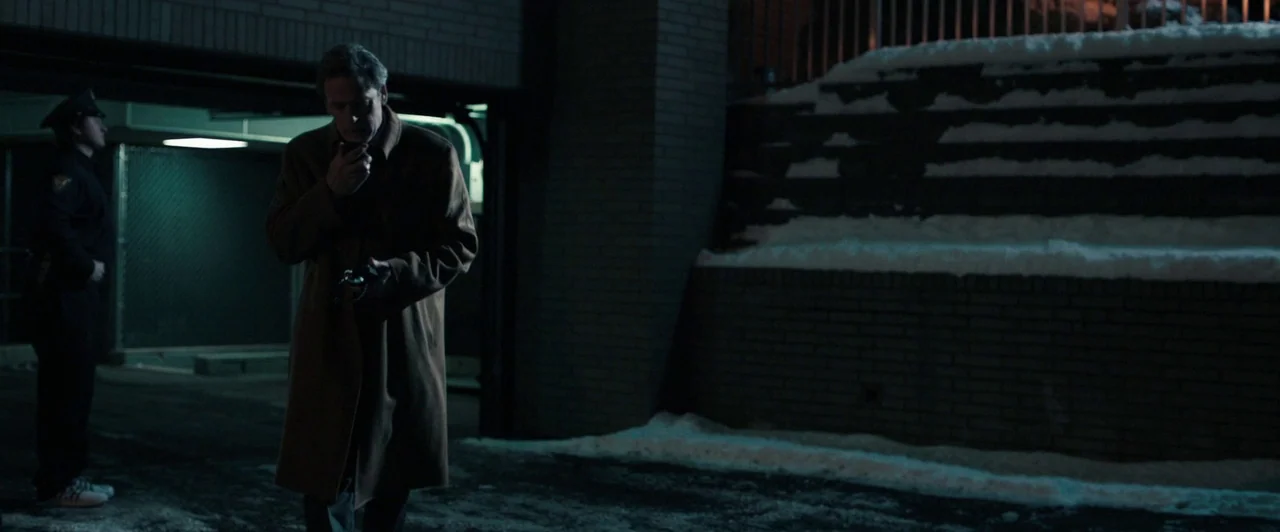 Compliance (2012) movie still: fullbody — A dark, snow-covered parking area next to a brick wall under fluorescent light. A man holds a radio while…