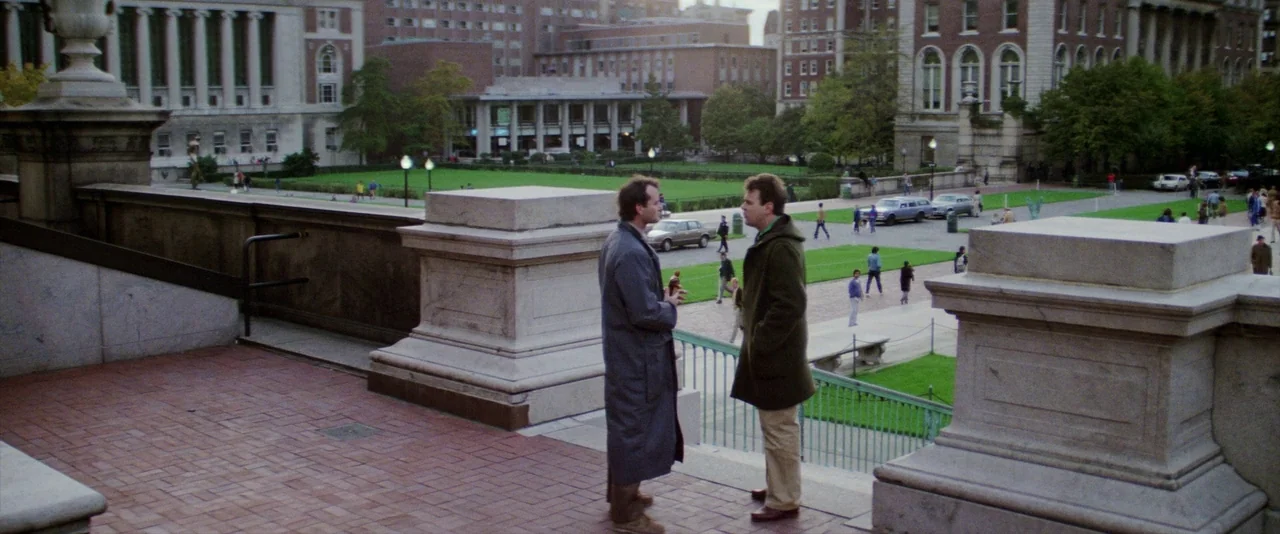 Ghostbusters (1984) movie still: fullbody — Two men talk on a stone balcony overlooking a large university campus quadrangle. Pedestrians walk on…
