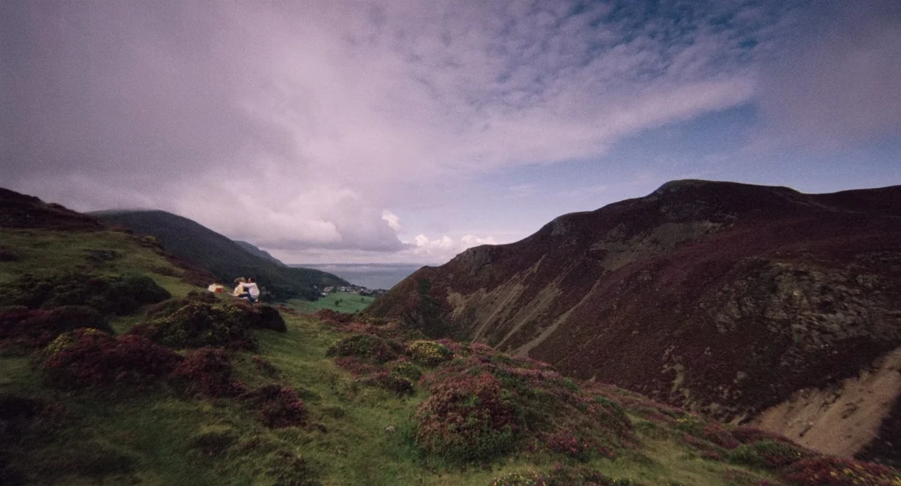 Highlander (1986) movie still: wide — A vast, rolling mountain landscape is covered in dark purple flowering shrubs and grass. Two small, indistinct…