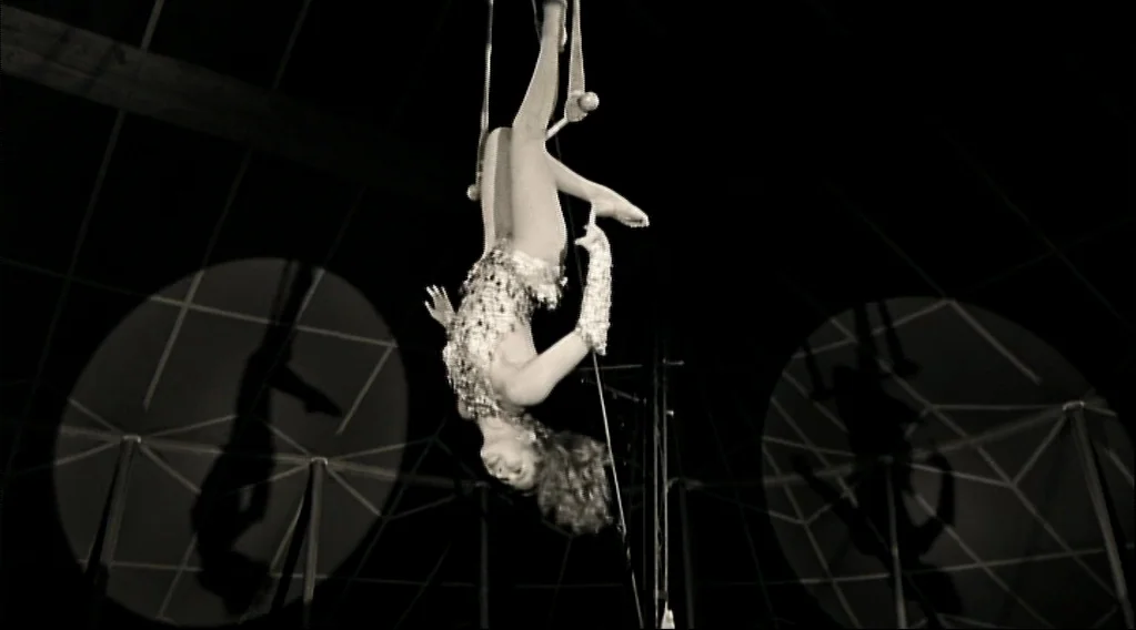 Wings Of Desire (1987) movie still: medium — The interior of a circus tent is shrouded in darkness behind a performer. A woman hangs inverted from a…