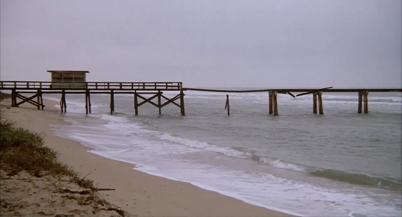 Empire of the Ants (1977) movie still: establishing — A long, damaged wooden pier stretches over the ocean under a grey overcast sky. The wooden…