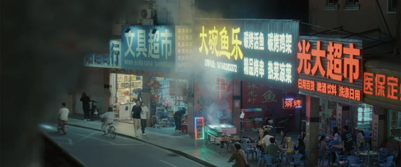 Yolo (2024) movie still: wide — A night view of a commercial city street with multiple glowing neon business signs. Pedestrians walk along the…