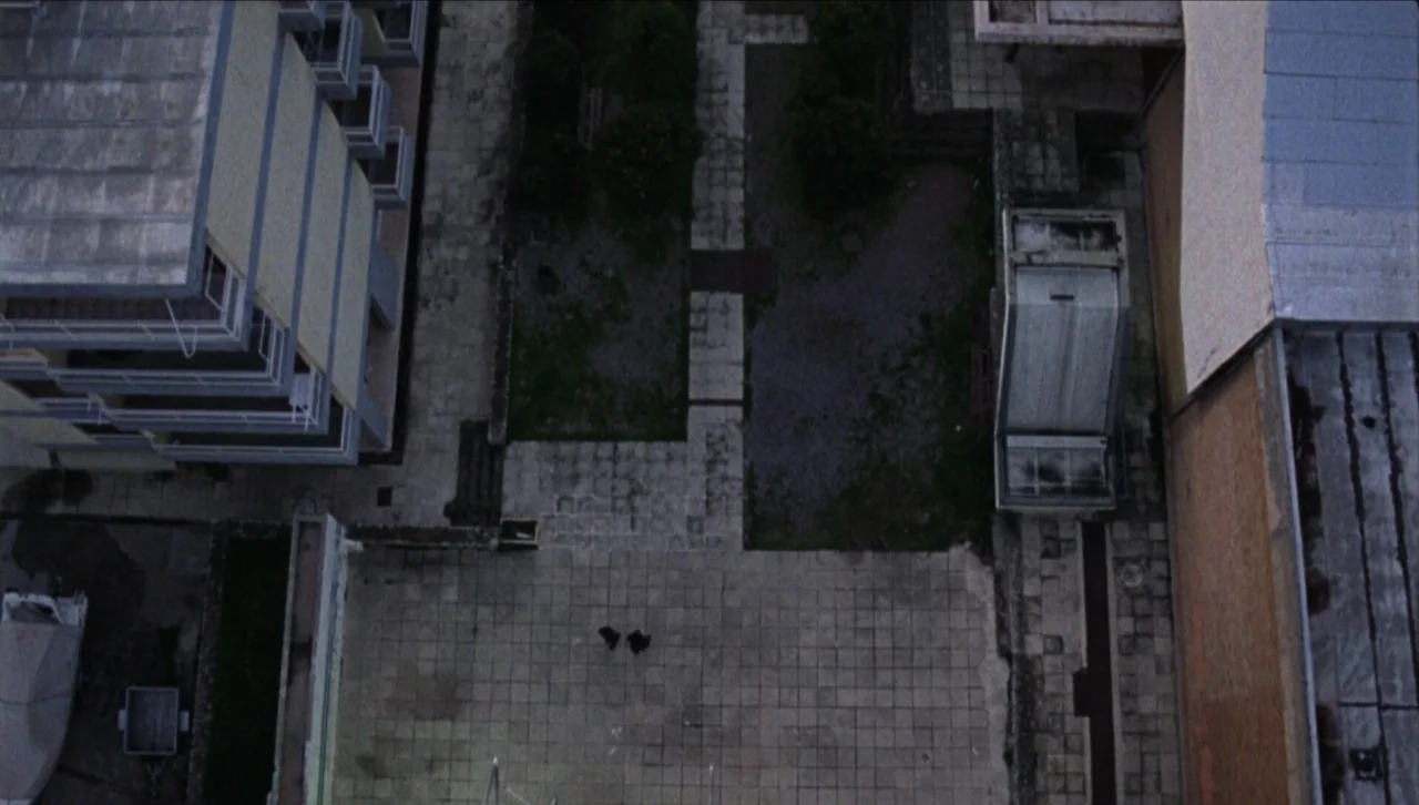 Attenberg (2010) movie still: establishing — An aerial view of a stone-paved courtyard nestled between two low-rise residential buildings. Two small…