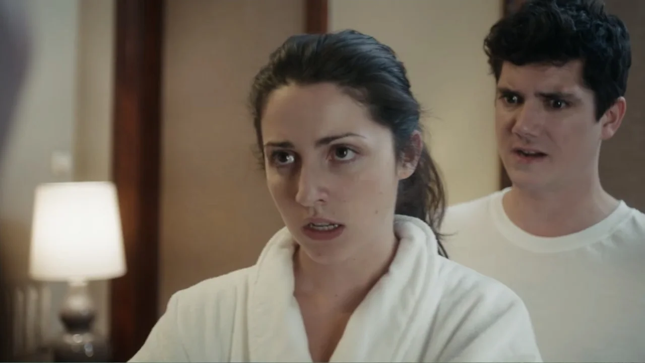 Toss It (2019) movie still: medium — An interior bedroom space with muted wall tones and a lamp in the background. A woman in a white bathrobe…
