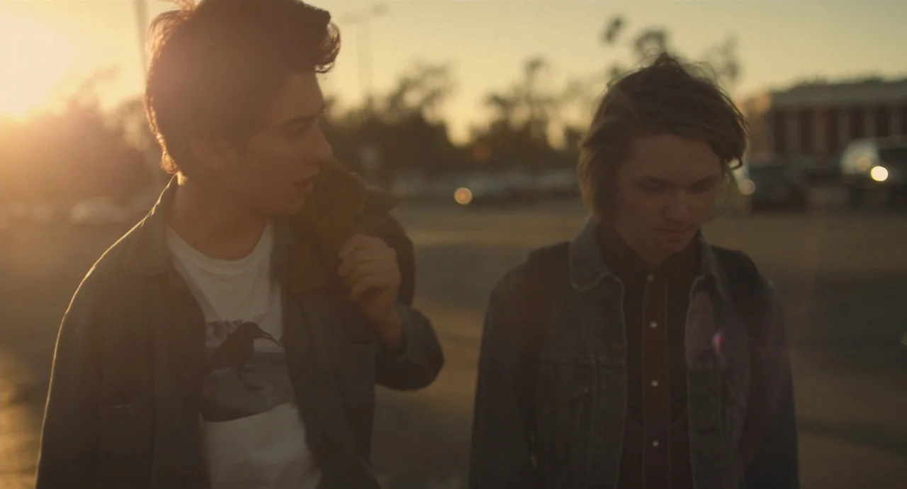 Palo Alto (2013) movie still: medium — A spacious paved parking lot is bathed in the warm light of golden hour. Two young men walk side by side…