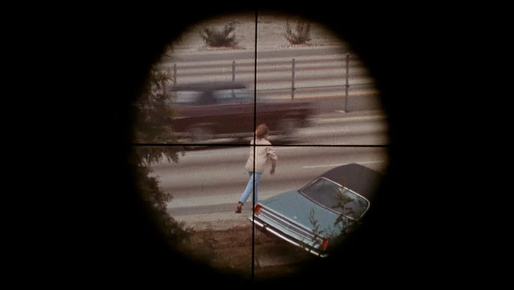 Targets (1968) movie still: wide — A view through a rifle scope shows a woman walking on the roadside. A blue vintage sedan is parked on the…