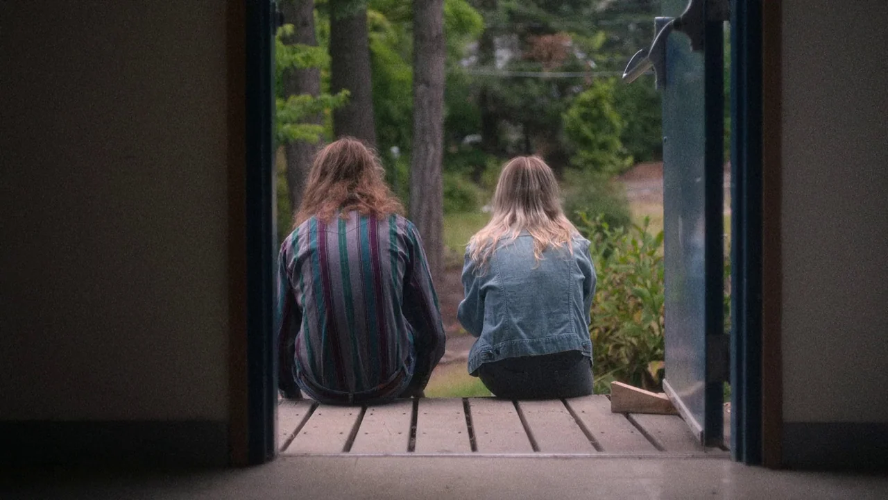 Showing Up (2022) movie still: medium — Two individuals sit on a wooden threshold within a dark framed doorway. They look out at a blurry treeline…