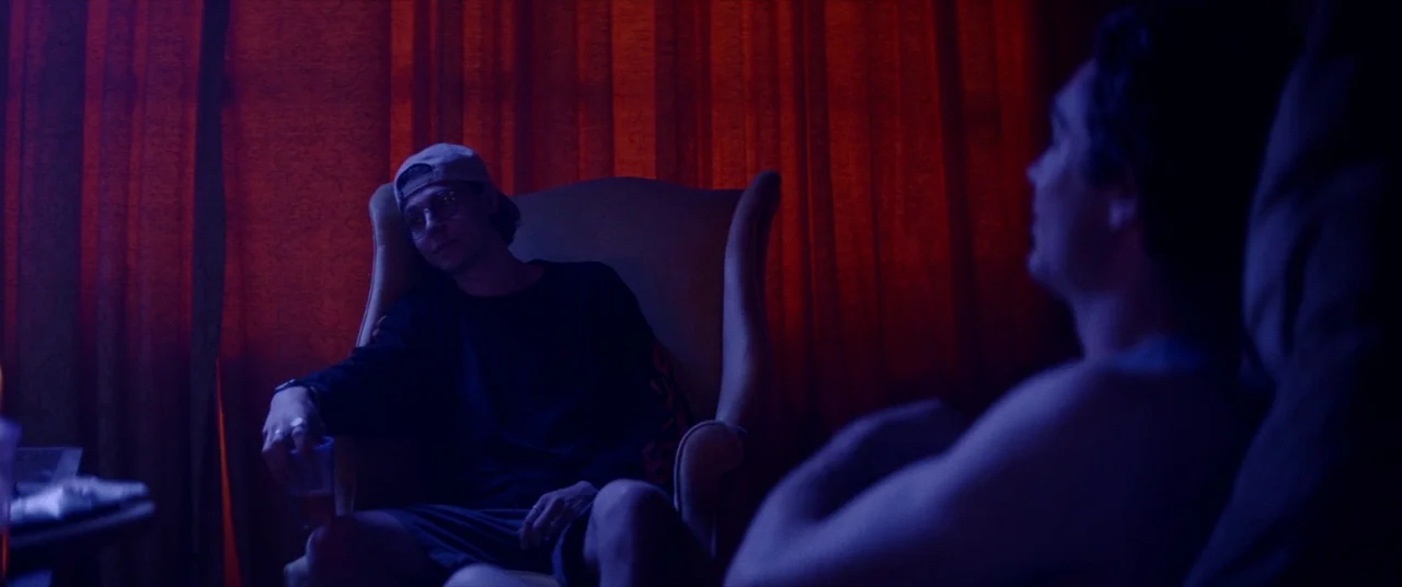 Assassination Nation (2018) movie still: medium — A dimly lit room with heavy red curtains hanging against a wall. Two young men sit in chairs…