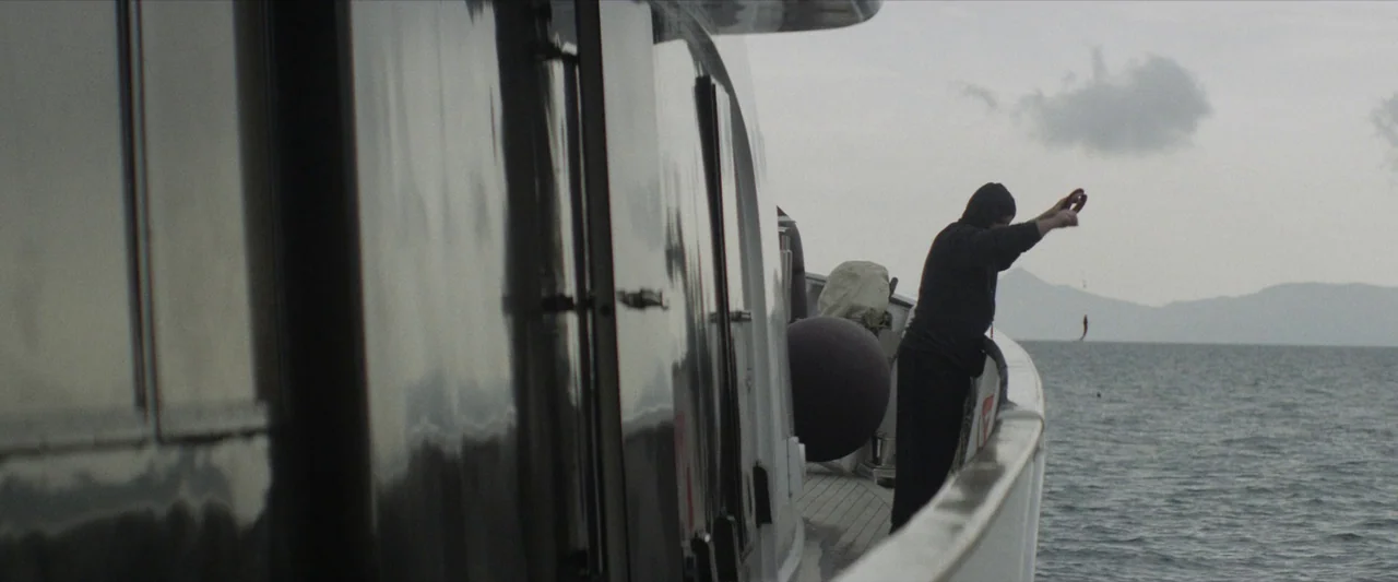 Chevalier (2015) movie still: wide — A person stands on the deck of a sleek boat near calm, open water. The individual holds a thin fishing line…