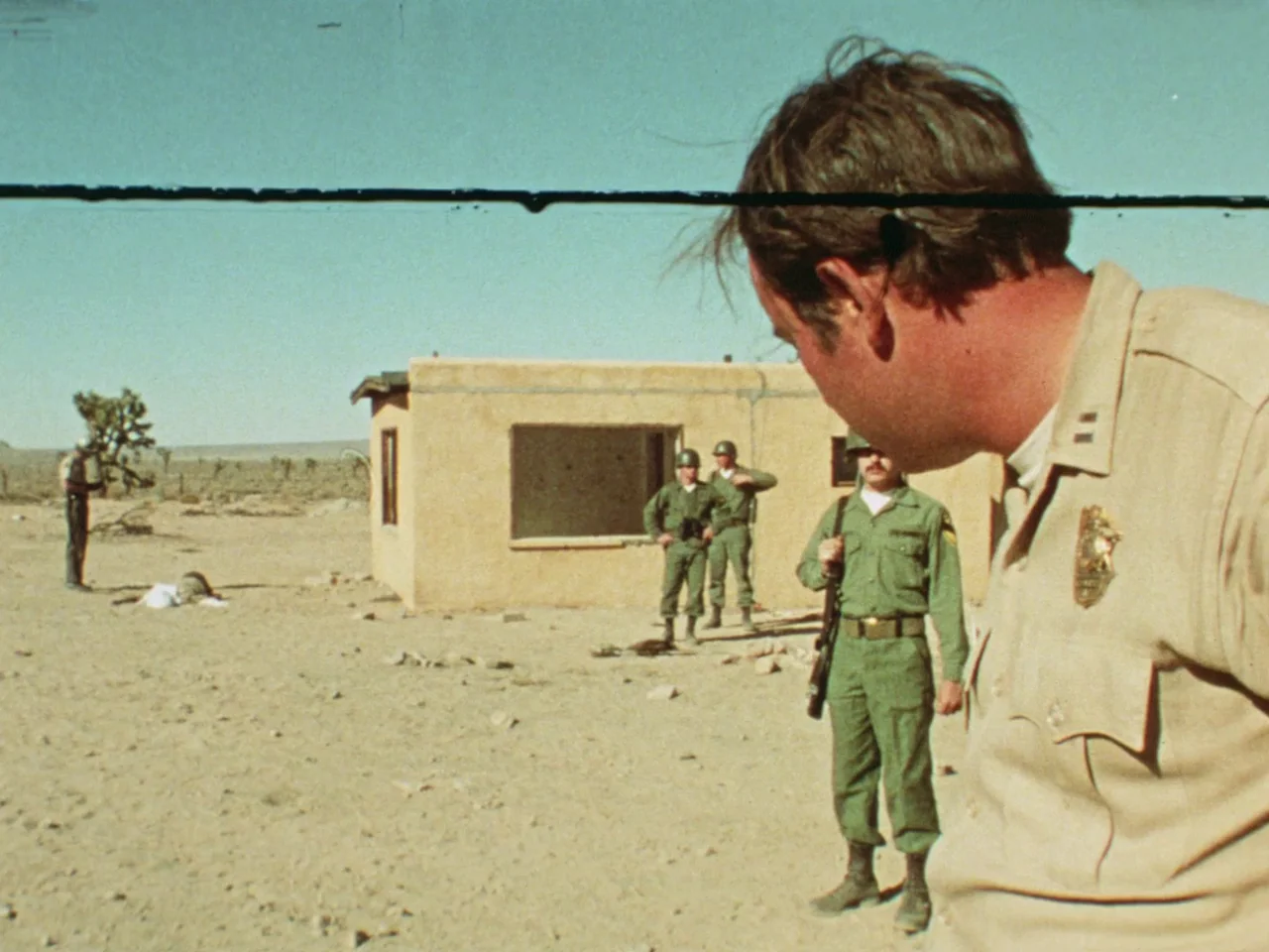 Punishment Park (1971) movie still: medium — A flat desert landscape features a small tan concrete building with open windows. Several armed…
