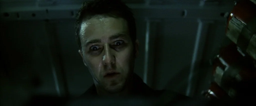 Fight Club (1999) movie still: closeup — The interior of a dark, cramped vehicle with ribbed structural metal walls. A man stares downward with…