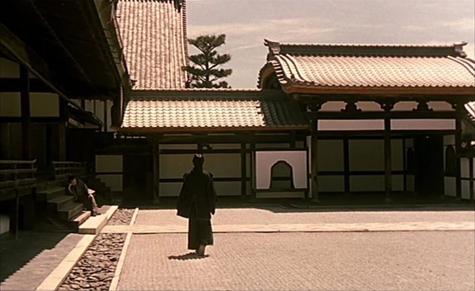 Gohatto (1999) movie still: wide — A traditional Japanese complex with dark timber framing and white plaster walls. A lone figure in dark clothing…