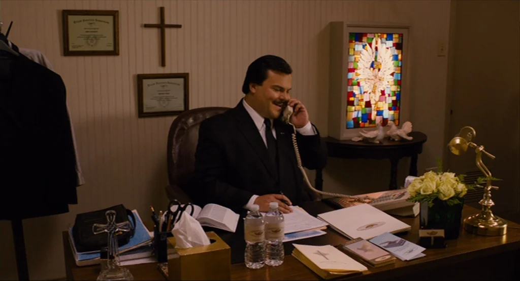 Bernie (2011) movie still: medium — A wood-paneled office contains a desk cluttered with documents, water bottles, and a telephone. A man in a dark…