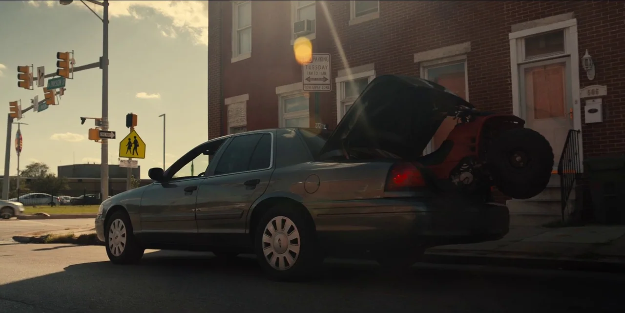 Charm City Kings (2020) movie still: wide — A grey sedan sits parked on a paved urban road near a red brick building. The car's trunk is open,…