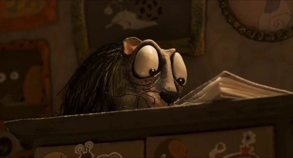 Memoir of a Snail (2024) movie still: closeup — A dimly lit room with framed artwork on a textured wall behind a desk. A puppet with large white…
